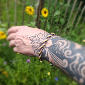 Op de afbeelding: Een gevlochten armband met paarse, gele en witte draden. De armband zit om een ​​getatoeëerde arm, met een vage achtergrond van zonnebloemen en een houten hek.