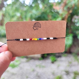 Könnte beinhalten: Ein handgefertigtes Armband mit einem Regenbogen-Perlen-Design. Das Armband hat schwarze, weiße, rote, orangefarbene, gelbe, grüne, blaue und violette Perlen. Das Armband wird auf einer braunen Karte mit einem Regenbogen-Herz-Logo präsentiert.