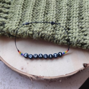 Könnte beinhalten: Ein schwarzes Armband aus Schnur mit dem Wort "EQUALITY" in weißen Buchstaben auf schwarzen Perlen. Die Perlen sind durch kleine regenbogenfarbene Perlen getrennt.
