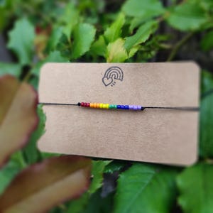 Könnte beinhalten: Ein Armband mit Regenbogenperlen auf einer braunen Karte mit einem Regenbogen- und Herzlogo. Das Armband hat kleine, farbige Perlen in einem Regenbogenmuster. Der Hintergrund ist verschwommenes grünes Laub.