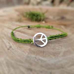 Könnte beinhalten: Ein grünes Makramee-Armband mit einem silbernen Friedenssymbol-Anhänger. Das Armband besteht aus gewebter grüner Kordel und hat ein kreisförmiges Friedenszeichen in der Mitte. Das Armband wird auf einer Holzoberfläche präsentiert.