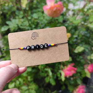 Könnte beinhalten: Ein handgefertigtes Armband mit schwarzen Buchstabenperlen, die "PRIDE" auf einer schwarzen Schnur bilden. Das Armband hat regenbogenfarbene Perlen auf beiden Seiten des Wortes. Das Armband wird auf einer kleinen braunen Karte mit einem Regenbogen-Logo präsentiert.