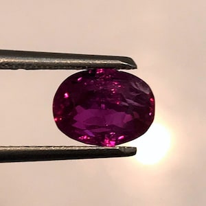 May include: A vibrant purple gemstone held by tweezers. The gemstone is round and has a smooth, polished surface.