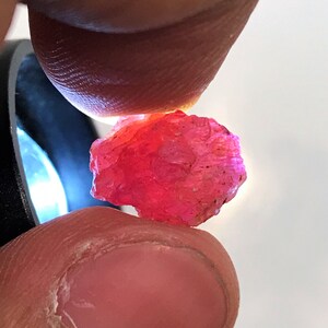 以下が含まれることがあります： ピンクでざらざらした不規則な形の宝石のクローズアップ。宝石は2本の指で挟まれています。