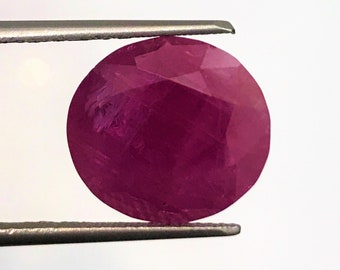 May include: A deep pink ruby gemstone with a round cut, held by tweezers against a white background.