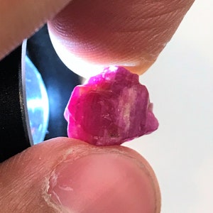 Pode incluir: Uma gema de rubi rosa vibrante com uma textura áspera e natural. O rubi é de uma cor rosa intensa e rica e tem uma aparência ligeiramente translúcida.