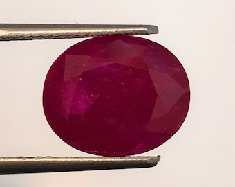 May include: A deep red ruby gemstone, with a round cut, is held by a pair of tweezers against a white background.
