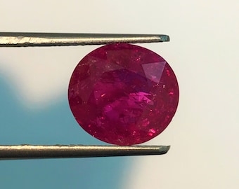 May include: A vibrant, deep red ruby gemstone held in tweezers. The ruby is round and has a smooth, polished surface.