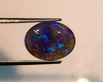 May include: An oval opal gemstone held by tweezers. The opal has a dark base colour with flashes of blue and green, and a hint of brown. The gemstone is polished and has a smooth surface, showcasing its natural beauty.