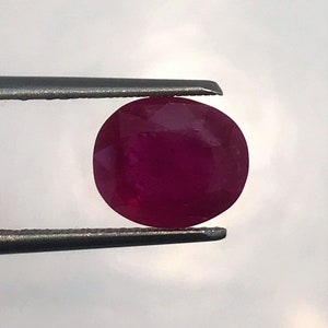 May include: A deep red oval-shaped gemstone held by tweezers.