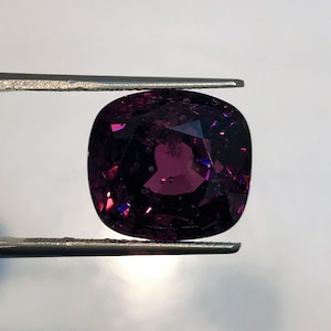 May include: A deep purple gemstone with a cushion cut, held by tweezers against a white background.