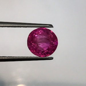 May include: A round, faceted, hot pink gemstone is held by a pair of tweezers. The gemstone has a polished surface and is a vibrant shade of magenta. The tweezers are made of metal and are holding the gem against a soft, blurred background.