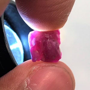 Könnte beinhalten: Nahaufnahme eines tiefrosa, facettierten Edelsteins, der zwischen zwei Fingern gehalten wird.