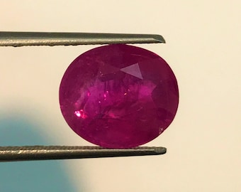 May include: A vibrant, oval-cut ruby gemstone with a deep magenta-red hue and multifaceted surface. The stone is held securely between the two metal tips of a pair of tweezers, showcasing its translucent quality and internal natural inclusions.