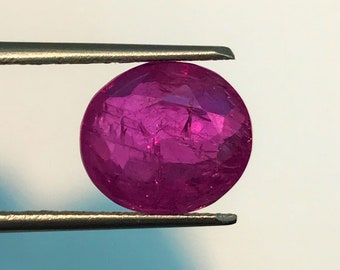 May include: A faceted, round, deep magenta gemstone is held by tweezers against a white background. The gem has internal inclusions and a polished surface, reflecting light. This gemstone is likely a ruby, a precious stone.