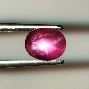 Puede incluir: Una piedra preciosa de rubí redonda y rosa con un reflejo en forma de estrella en el centro.