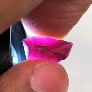 Puede incluir: Una piedra preciosa facetada de color rosa intenso con un acabado rugoso y natural. La piedra preciosa se sostiene en una mano, mostrando su tamaño y forma.