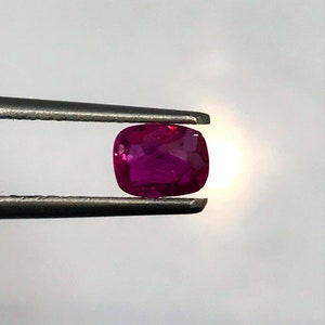 May include: A vibrant, deep red gemstone with a cushion cut. The gemstone is held in place by a pair of tweezers.