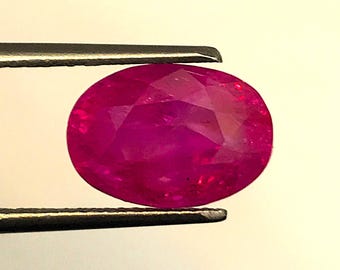 May include: A faceted, oval ruby gemstone held by tweezers. The ruby is a deep, vibrant red colour, with multiple facets that catch the light. The tweezers are silver-coloured.