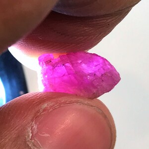 May include: A vibrant pink, faceted gemstone with a rough, natural texture. The gemstone is held between two fingers.