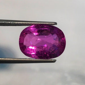 Puede incluir: Una gema ovalada de color rosa vibrante sostenida por unas pinzas. La gema tiene una superficie lisa y pulida y un ligero brillo.