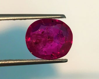 May include: A vibrant pink oval-shaped gemstone held by tweezers. The gemstone has a slightly cloudy appearance with a few small inclusions.