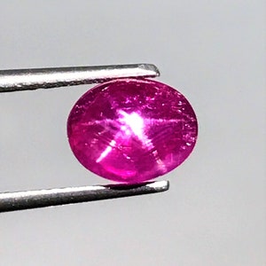 Può includere: Una pietra preziosa di rubino stellato rotonda, rosa, con una stella a sei punte al centro. Il rubino è tenuto da una pinzetta.