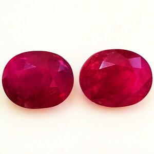 Puede incluir: Dos piedras preciosas ovaladas y facetadas de color rojo intenso. Las gemas se muestran sobre un fondo blanco, mostrando sus superficies pulidas y su claridad interna. Las piedras son de tamaño y forma similares, lo que sugiere que son un par a juego.