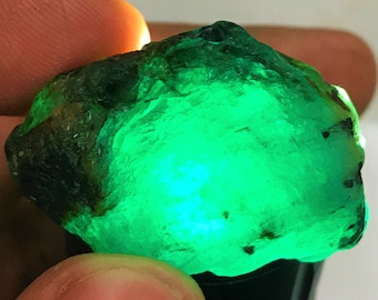 May include: A large, rough, green emerald gemstone with a translucent, glassy appearance. The emerald is illuminated from below, highlighting its vibrant green color.
