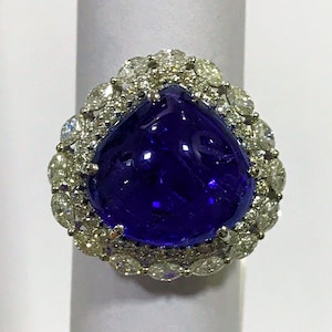 May include: A pear-shaped blue gemstone ring with a halo of smaller diamonds. The ring is set in white metal.