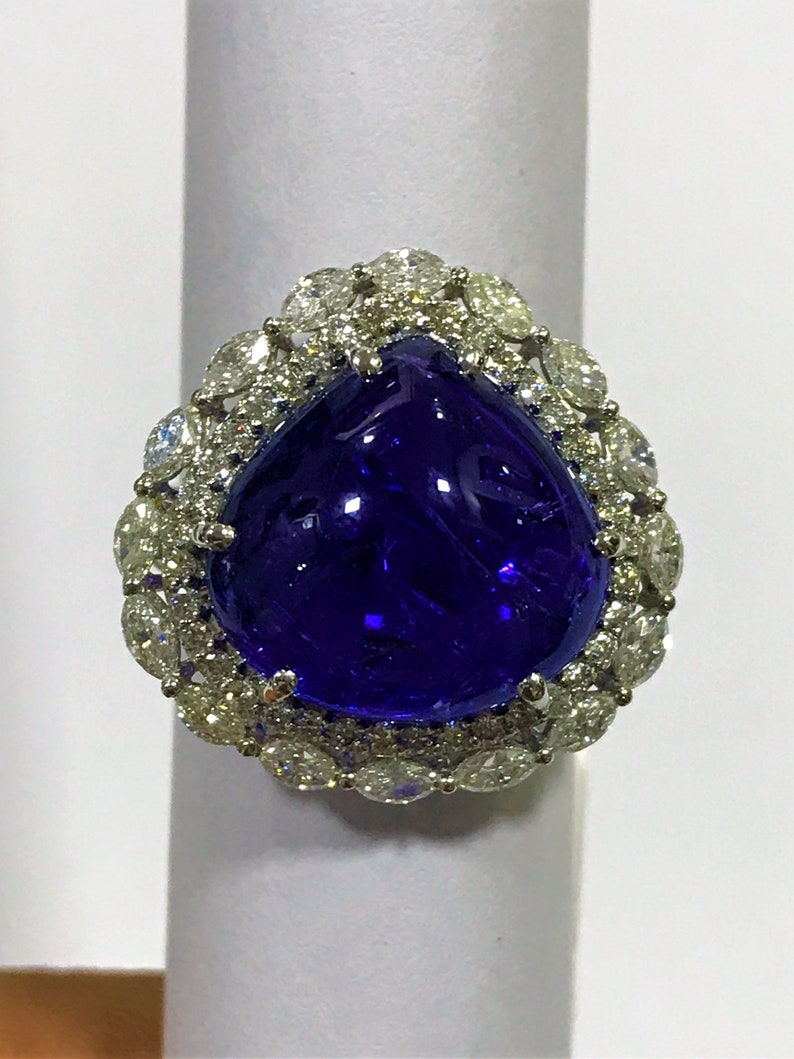 May include: A pear-shaped blue gemstone ring with a halo of smaller diamonds. The ring is set in white metal.