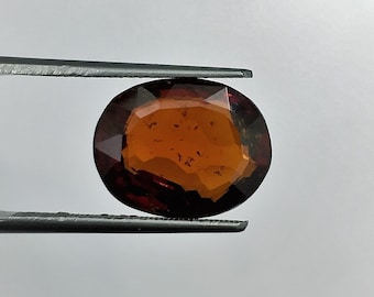 May include: A close-up of a brown oval gemstone held by tweezers. The gemstone has a reddish-brown hue and a faceted cut.