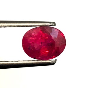 Op de afbeelding: Een ronde, rode edelsteen met een glad, gepolijst oppervlak. De edelsteen wordt vastgehouden in een pincet.