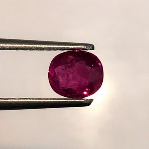 Puede incluir: Una gema de rubí ovalada y vibrante sostenida en pinzas. El rubí es de color rojo intenso con un ligero tono púrpura.