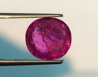 May include: A vibrant pink oval-shaped gemstone held by tweezers. The gemstone has a smooth, polished surface and a slightly translucent appearance.