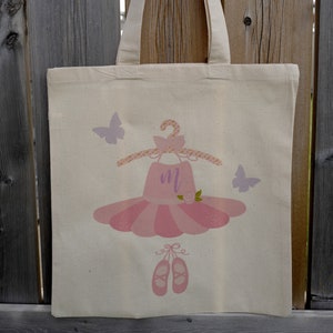 Peut inclure: Un sac fourre-tout en toile blanche avec un motif de robe de ballerine rose et de chaussures. La robe a un nœud violet et la lettre "M" sur le corsage. Il y a deux papillons violets sur le sac.