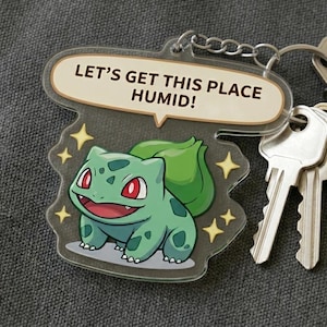 May include: A clear acrylic keychain featuring a green Bulbasaur character with red eyes and the text "LET'S GET THIS PLACE HUMID!". The keychain is attached to a silver keyring with keys.