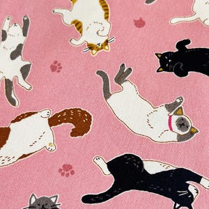 May include: Pink fabric with a repeating pattern of playful cats in various poses. The cats are in shades of black, white, brown, and gray. Scattered paw prints add to the whimsical design. The fabric is folded, showing its texture and the pattern's continuity.