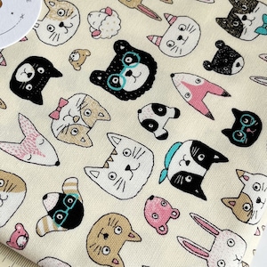 May include: A cream-colored fabric with a repeating pattern of black, brown, white, and pink animal faces. The fabric features cats, rabbits, bears, and other animals. The fabric is 13 inches wide.