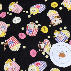 May include: Black fabric with a repeating pattern of cartoon cats holding boxes of popcorn. The boxes are red, white, and blue with the words "Pop Corn" on them. The cats are wearing different expressions and some have speech bubbles with words like "Hungry?" and "Caramel".