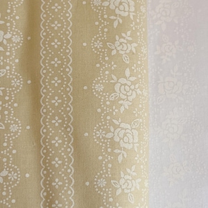 May include: A beige fabric with a white floral pattern. The fabric has a repeating pattern of small white flowers and a larger white rose with leaves. The fabric is folded in half and the pattern is visible on both sides.