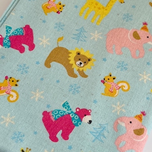 Könnte beinhalten: Ein hellblauer Stoff mit einem sich wiederholenden Muster von Tieren, die Wintermützen und Schals tragen. Zu den Tieren gehören Bären, Giraffen, Elefanten und Löwen. Der Stoff ist perfekt für Bastelprojekte wie Quilts, Decken und Kissen.