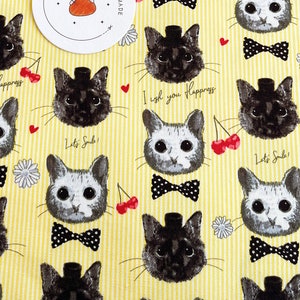 May include: Yellow fabric with a repeating pattern of black and white cats wearing bow ties. The cats have different facial expressions and some have a top hat. The fabric has a white stripe pattern and red cherries and white flowers are scattered throughout. The text "I wish you happiness" and "Let's Sub" are repeated throughout the fabric.