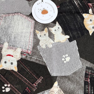 May include: A black and gray fabric with a repeating pattern of denim pockets and cartoon cats. The cats are in various poses, some peeking out of the pockets, some sitting inside, and some lying down. The fabric has a distressed look, with faded areas and a slightly rough texture.