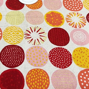 May include: A roll of fabric with a repeating pattern of large, colorful circles in shades of red, yellow, orange, and pink on a cream background. A wooden ruler with inch markings and a small sticker are also visible.