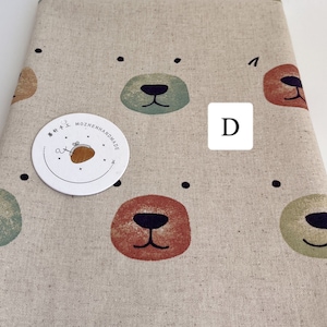 May include: Beige fabric with a repeating pattern of colourful bear faces. The fabric has black dots scattered throughout. A white circle label with the text "MOZHENHANDMADE" is attached to the fabric. The letter "D" is written in black on a white square.