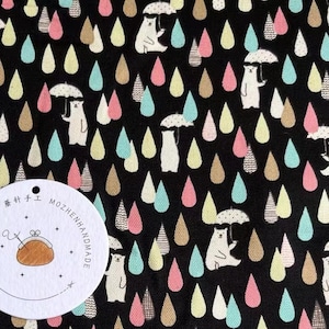 Op de afbeelding: Zwarte stof met een herhalend patroon van witte beren die paraplu's vasthouden en kleurrijke regendruppels. De stof heeft een label met de tekst "MOZHENHANDMADE" en "85".