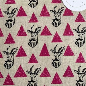 Pode incluir: Um tecido bege com um padrão repetido de triângulos rosa e ilustrações em preto e branco de uma cabra com barba e bigode. O tecido tem uma etiqueta com o texto "MOZHENHANDMADE" em preto e branco.