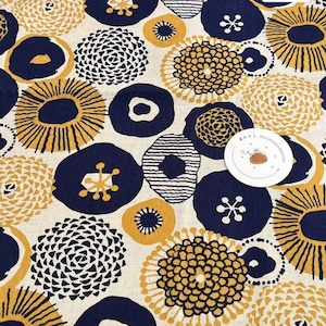 Könnte beinhalten: Ein beigefarbener Stoff mit einem sich wiederholenden Muster aus marineblauen und goldenen Kreisen. Die Kreise haben verschiedene Designs, darunter ein Sonnenstrahlenmuster, ein Wirbelmuster und ein einfacher Kreis. Der Stoff ist mit "Project B" und "Cafe de Cotton" gekennzeichnet.