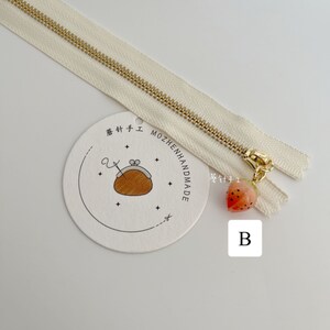 May include: A white zipper with a gold zipper pull. The zipper pull has a small strawberry charm attached to it. The zipper is on a white background with a round tag that says "Mozhenhandmade" in Japanese and English. The tag also has a small illustration of a purse with a needle and thread.